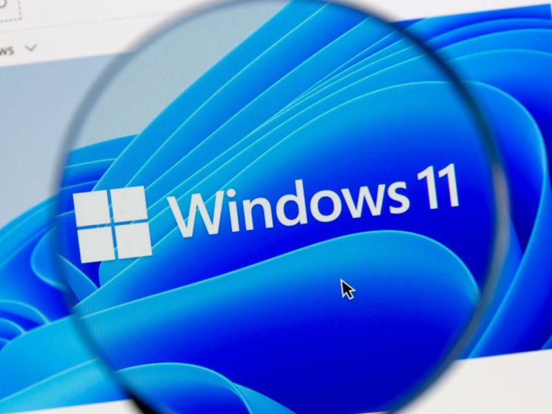 Windows 11-Logo auf einem Bildschirm. Eine Lupe vergrößert es zusätzlich.